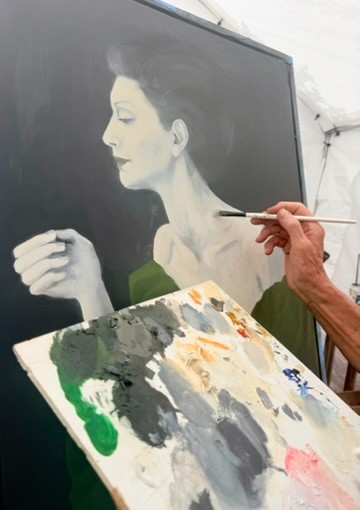 Janet's hand raising a fine brush to a large monochrome portrait in progress, a loaded painter's palette of greens, greys and ochre in the foreground.
