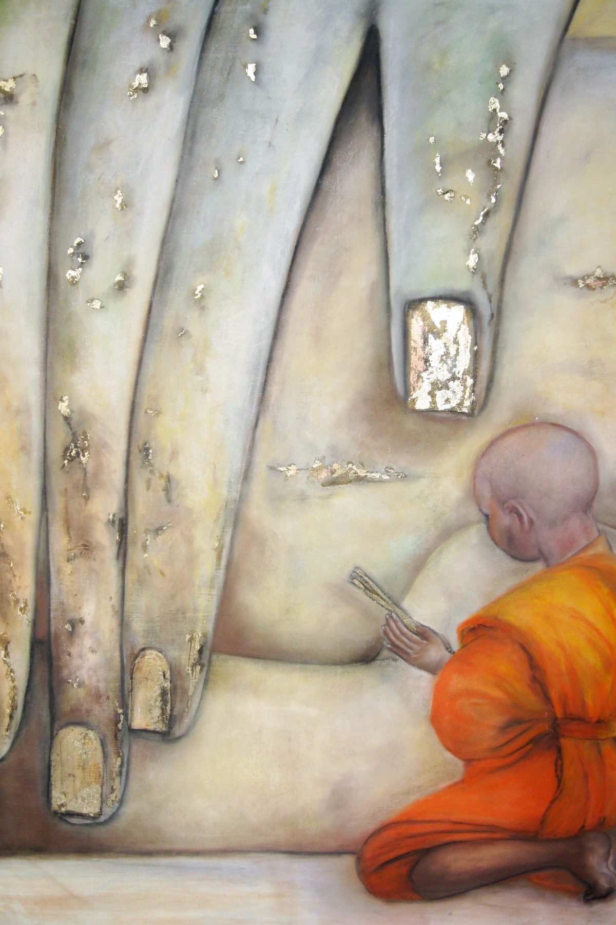 A small robed monk in saffron kneeling and reading at the feet of towering gold-leafed figures.