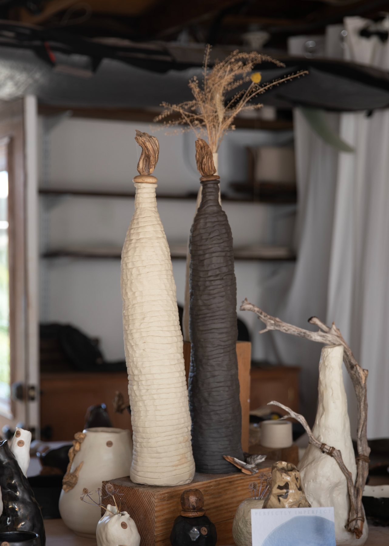 Two tall bottle vases, one cream, one charcoal, with deeply ridged surfaces and driftwood stoppers, standing on a wooden plinth in the studio.