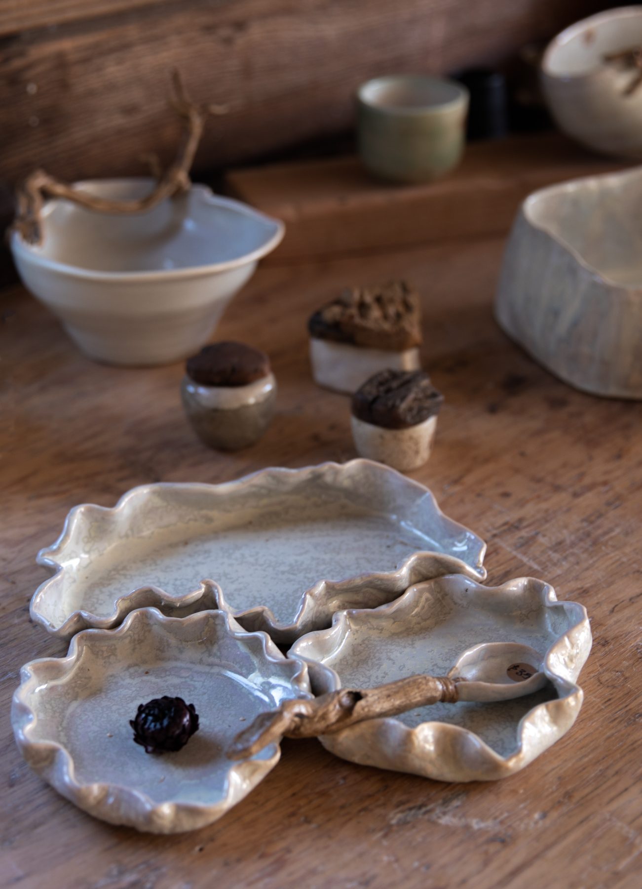 Handmade ceramic dishes and vessels with rippled edges and driftwood handles, arranged on a worn wooden table.
