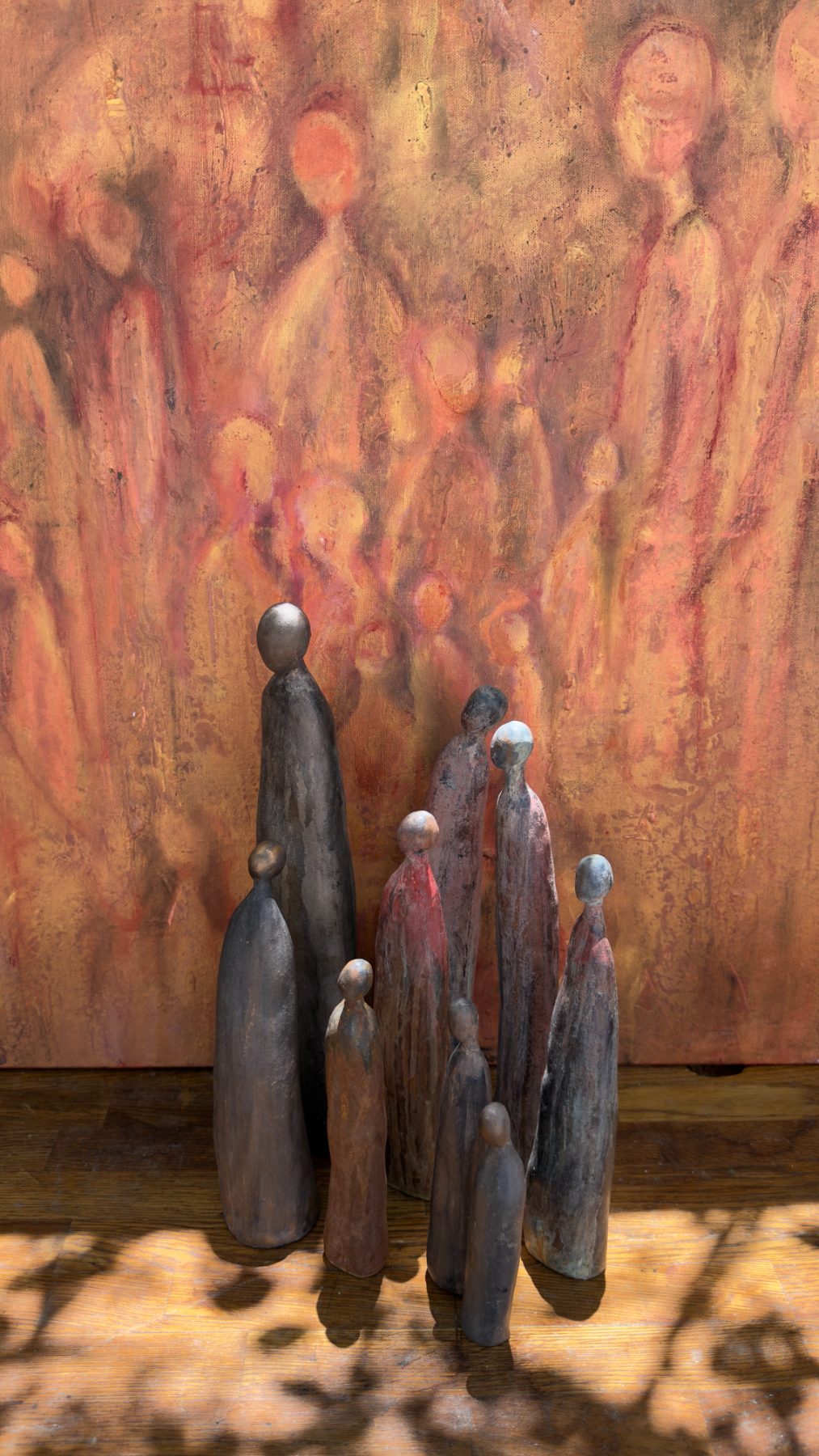 A gathering of tall, slender ceramic figures standing close together on a wooden floor, set against a painting of luminous rose-gold figures.