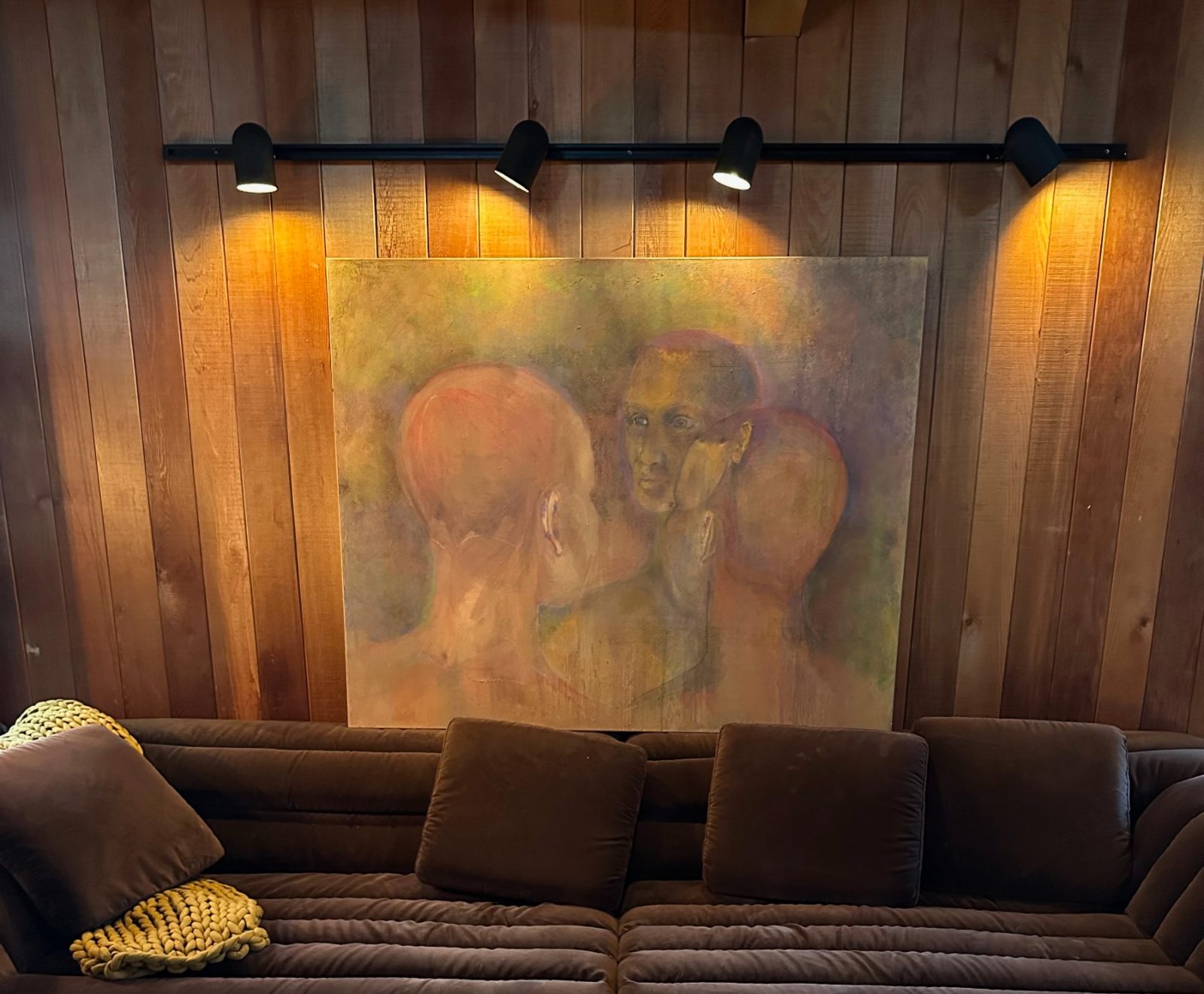 A large original Janet Fryer painting of three heads hanging above a sofa in a warm wood-panelled room, lit by picture lights.