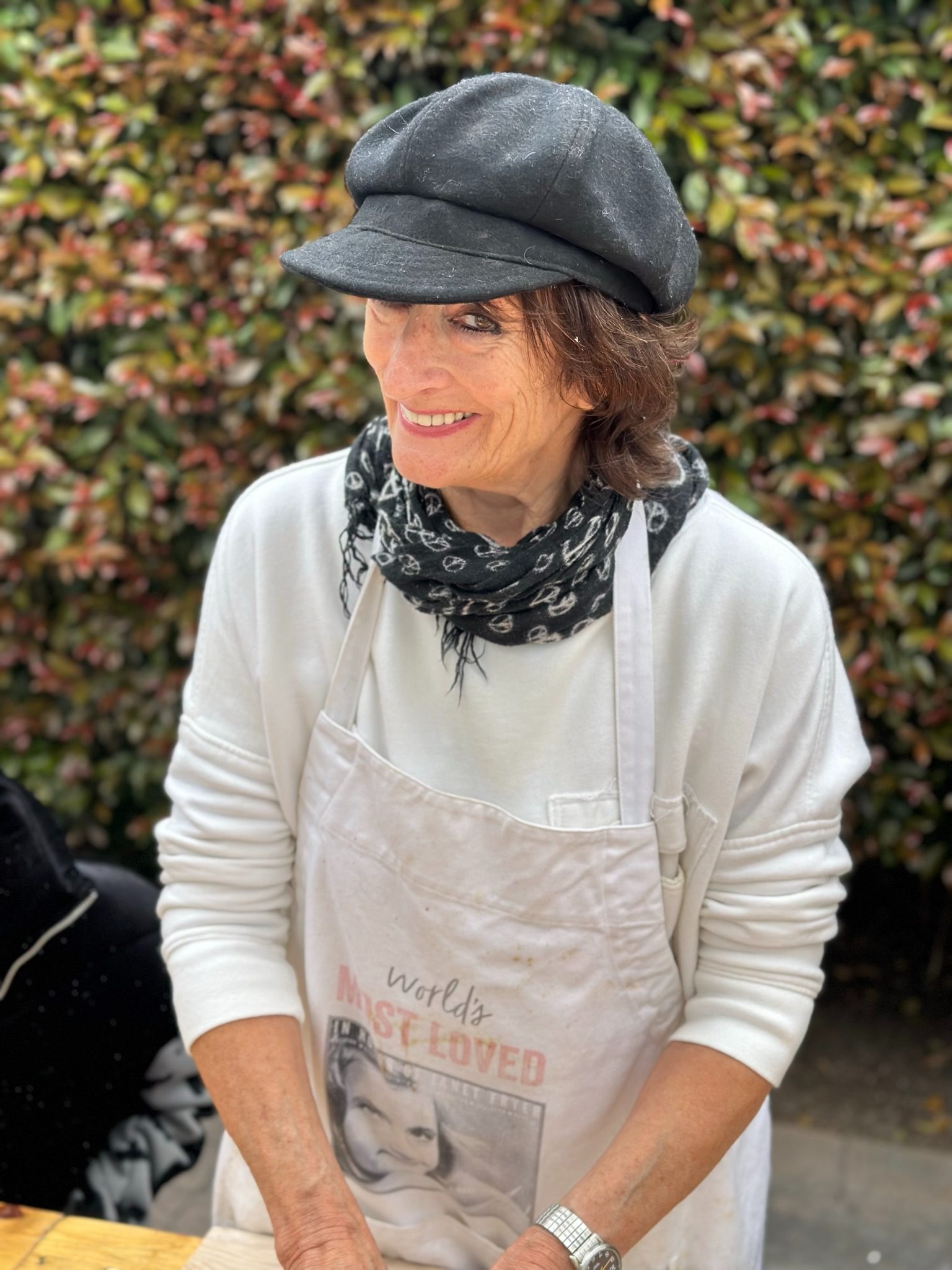 Photograph of Janet Fryer outdoors in a dark cap, patterned scarf and paint-marked apron, smiling and looking to the side against an autumn hedge.