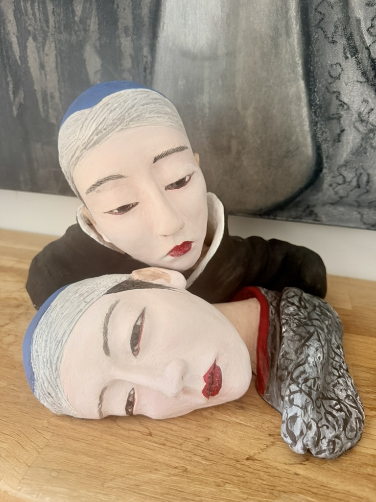 The clay version of The Gieshas, two sculpted faces laid on a pale surface, one figure resting its head against the other, painted in white, black, blue and red.