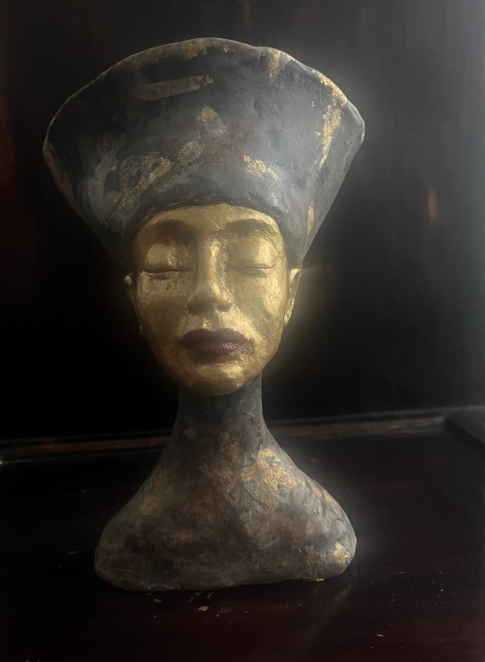 A sculpted head of a goddess with closed eyes and serene face, gold leafed skin and rose lips, wearing a tall dark headdress smudged with bronze, on a shaped torso plinth.