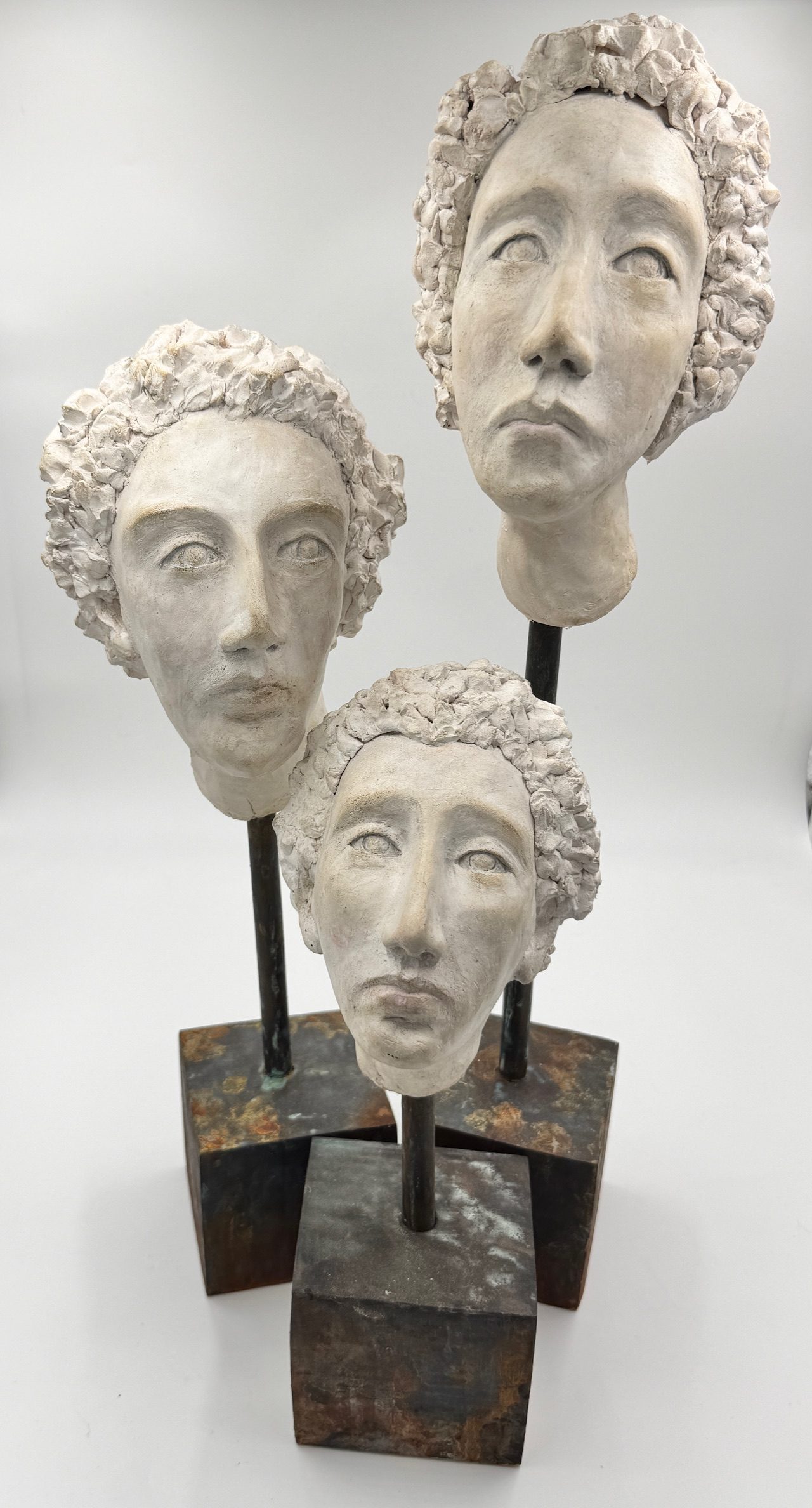 Three sculpted clay faces raised on dowels set in solid wood blocks, arranged as a triad, each with an aquiline nose, firm mouth and tightly curled hair, painted in aged white.
