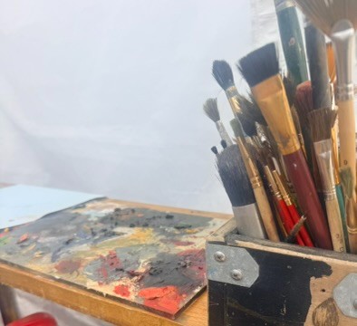 Janet's studio worktable, a well-used palette thick with mixed colour beside a jar of brushes, in warm daylight.