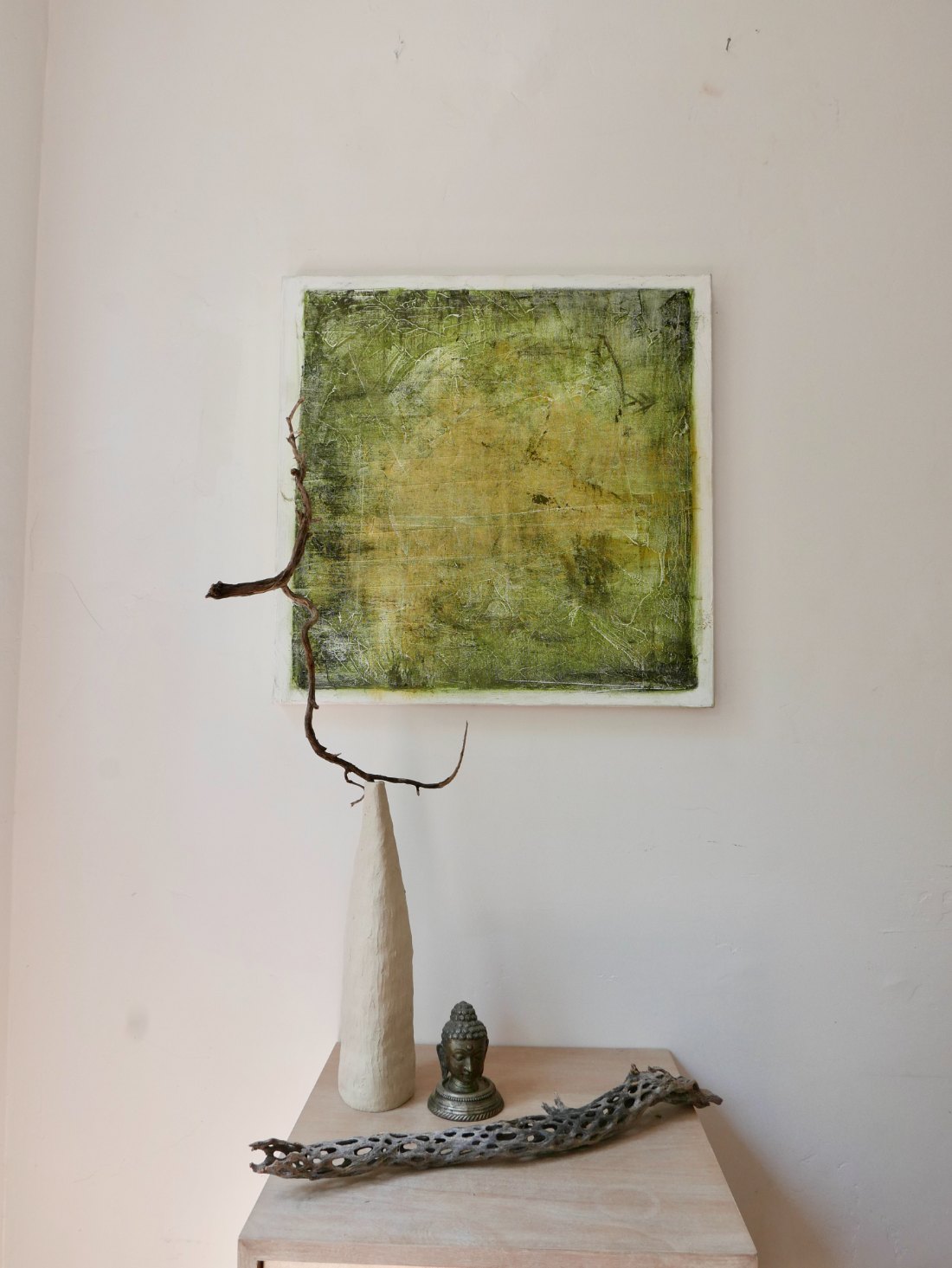 A green and gold abstract canvas hanging above a console with a pale ceramic vessel, driftwood and a small bronze Buddha.