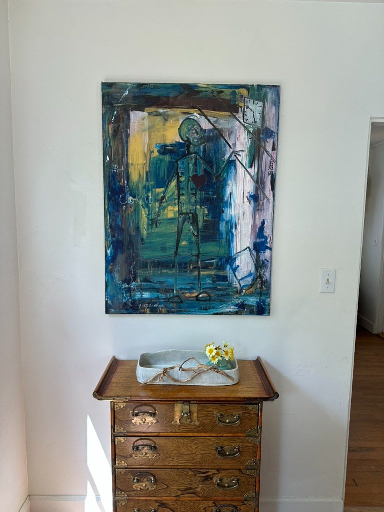A blue-toned painting of a figure holding a red heart, hanging above an antique wooden chest with fresh daisies.