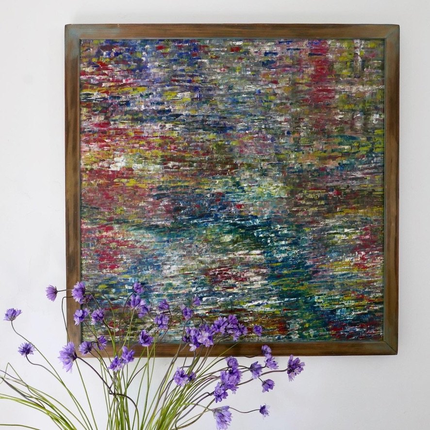 A densely coloured, framed mosaic-like painting on a white wall with purple wildflowers rising in front of it.